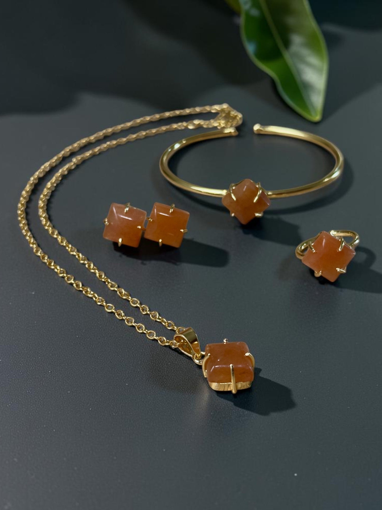 Gold jewelry set featuring a necklace, cuff bracelet, and stud earrings, each adorned with a faceted orange gemstone, displayed on a dark surface with green leaves in the background.