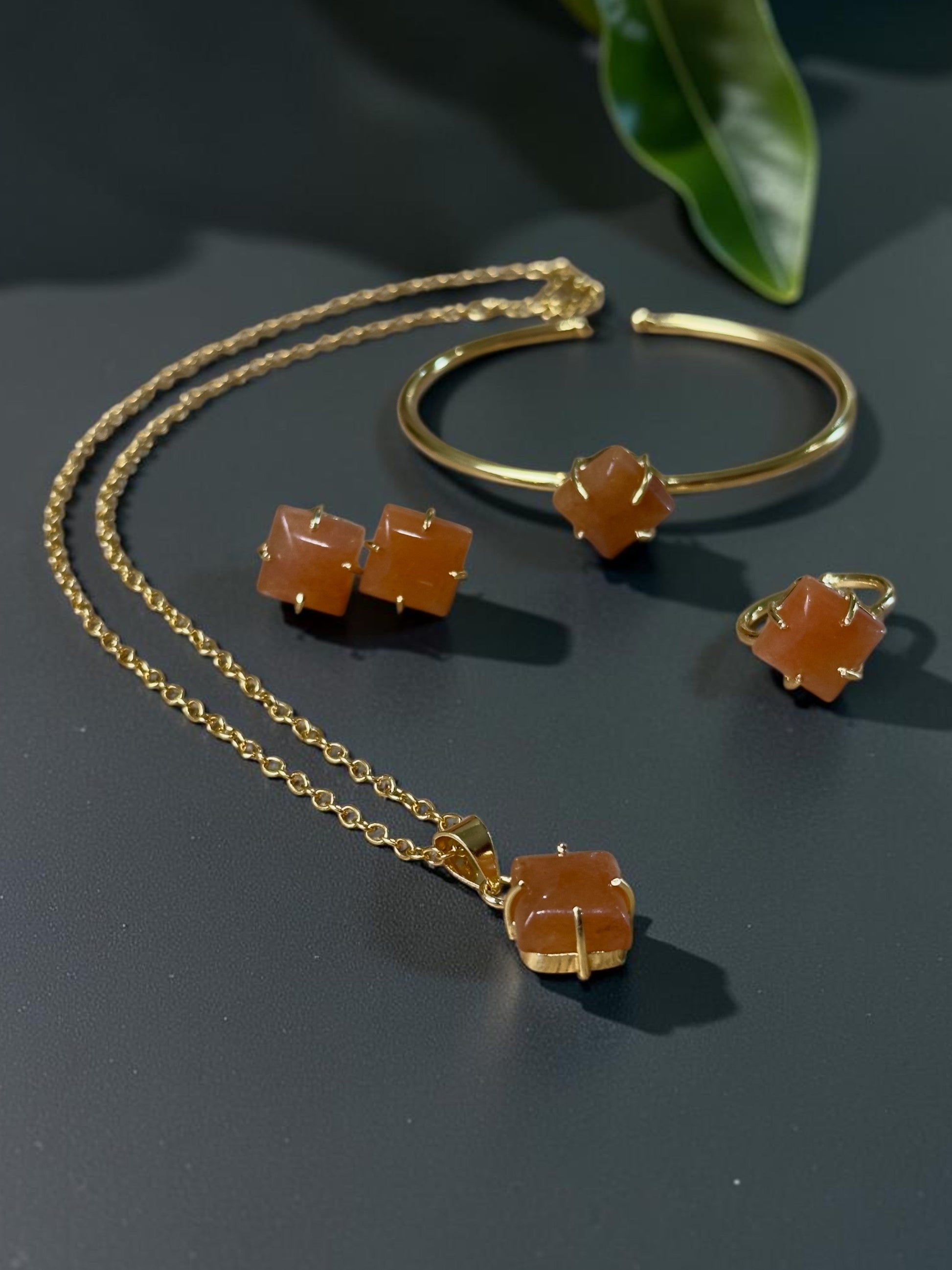 Gold jewelry set featuring a necklace, cuff bracelet, and stud earrings, each adorned with a faceted orange gemstone, displayed on a dark surface with green leaves in the background.