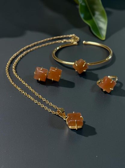 Gold jewelry set featuring a necklace, cuff bracelet, and stud earrings, each adorned with a faceted orange gemstone, displayed on a dark surface with green leaves in the background.